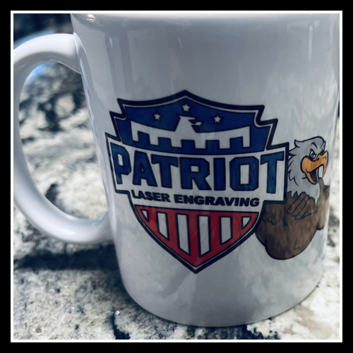 A photograph of a white 15 ounce ceramic mug on a table with the Patriot Laser Engraving Logo sub laminated on it.